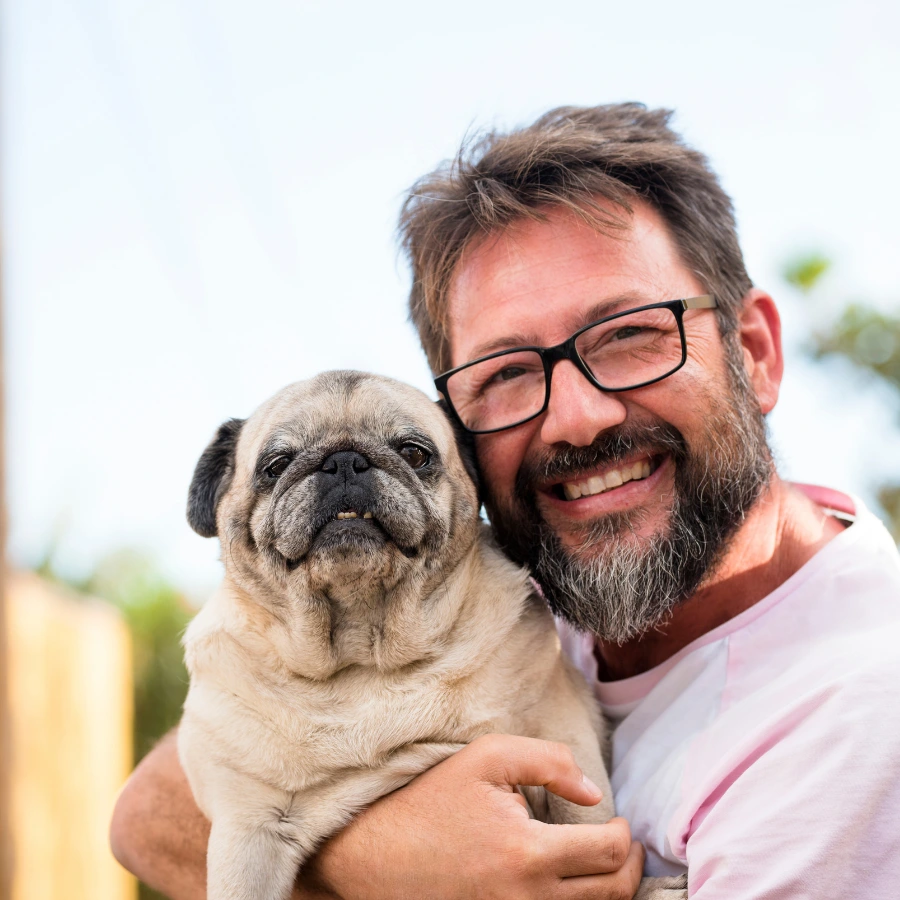 Smiling man with black glasses holding a fawn-colored pug close to his face outdoors in soft daylight.