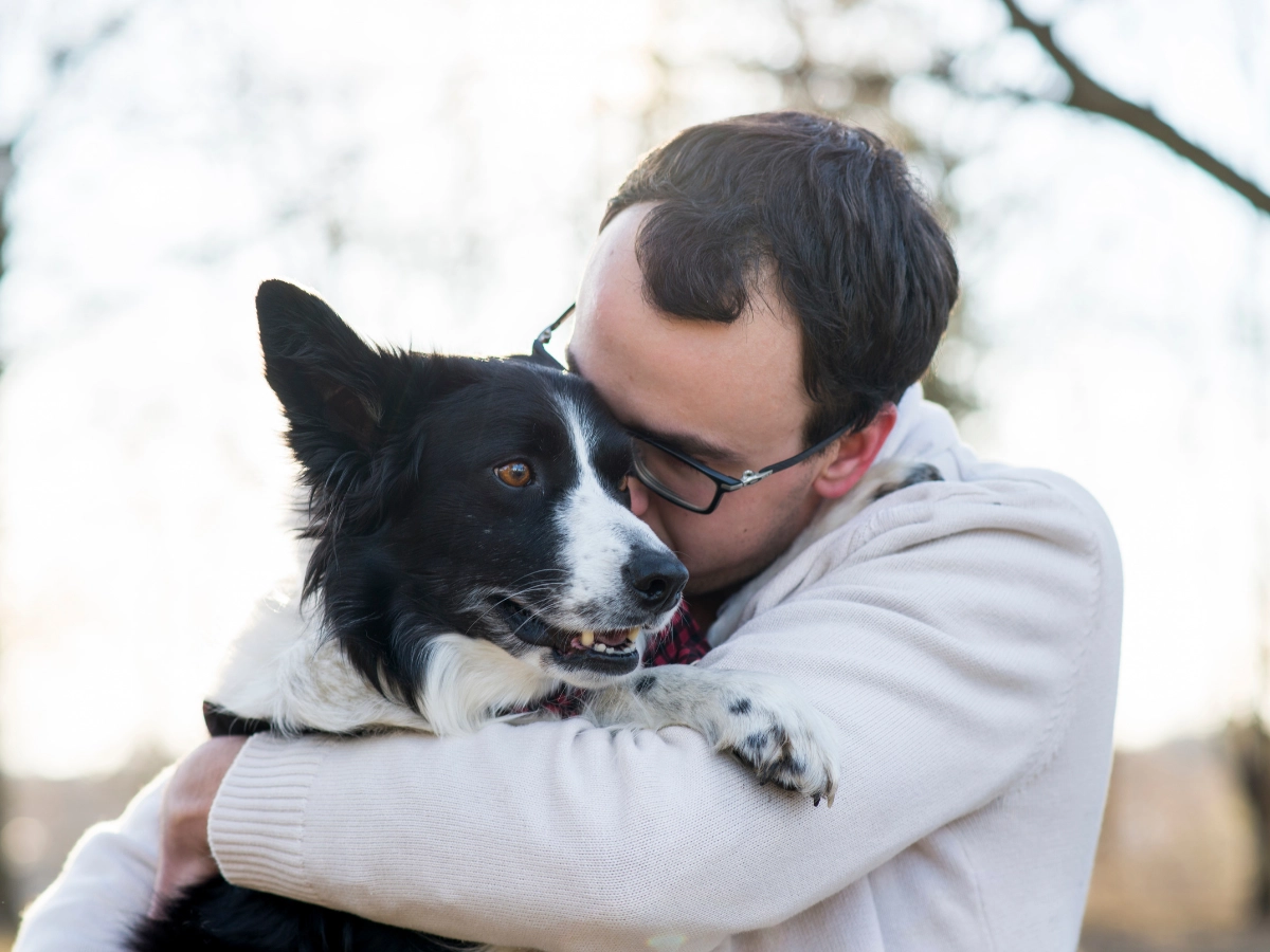 A person in a light hoodie hugs a black-and-white dog with one paw on their arm outdoors in soft, snowy light.