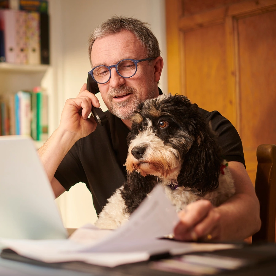 A man in glasses holds a phone to his ear while sitting at a table with a small dog in his arms, looking toward the camera.