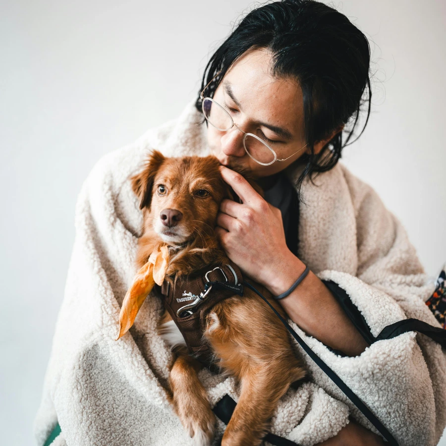 A person in a light robe cuddles a small brown dog while gently holding its head, with the dog looking toward the camera against a plain light background.