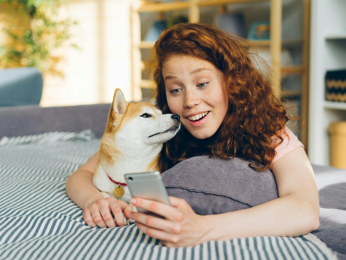 A woman smiling while cuddling a white-and-tan dog on a bed, both looking at a smartphone in her hands in a bright living room.