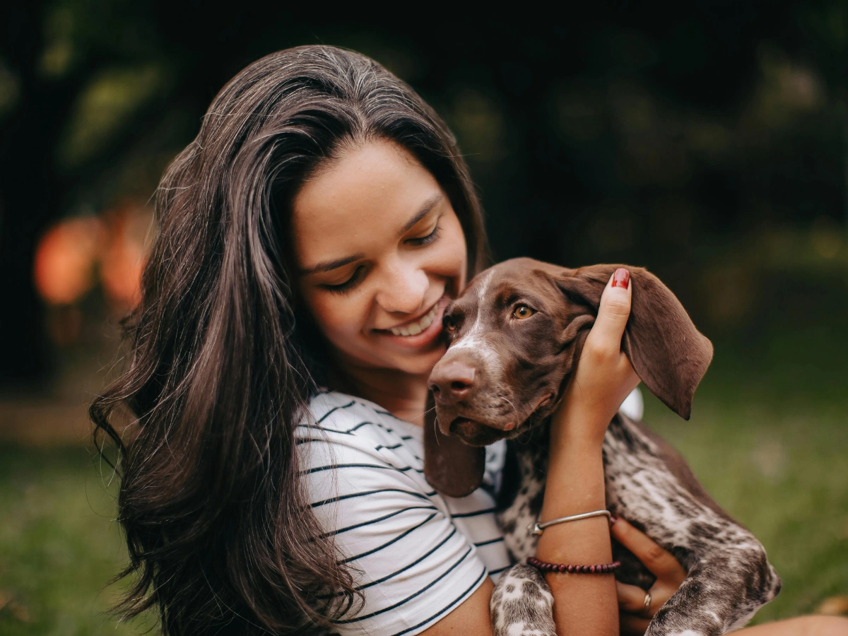 A person smiling while gently holding and stroking a brown-and-white dog outdoors at dusk, with a soft blurred green background and warm lighting.