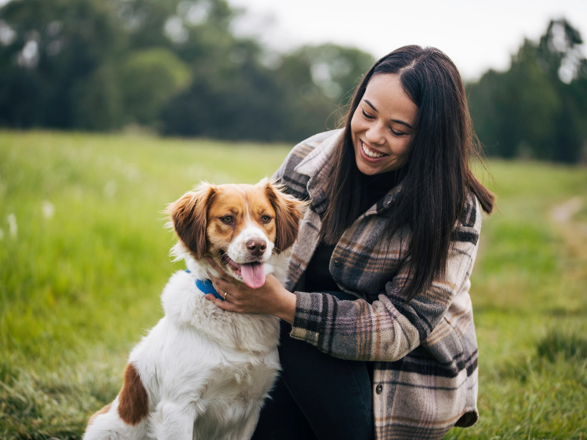 A woman in a plaid jacket hugs a friendly dog with a blue collar while sitting on green grass in a sunny park, both smiling.