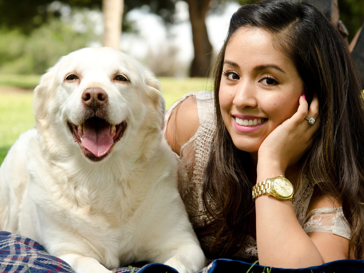 Woman smiling outdoors while resting her head on her hand beside a happy white dog with its mouth open, both seated on a blanket in a sunny park.