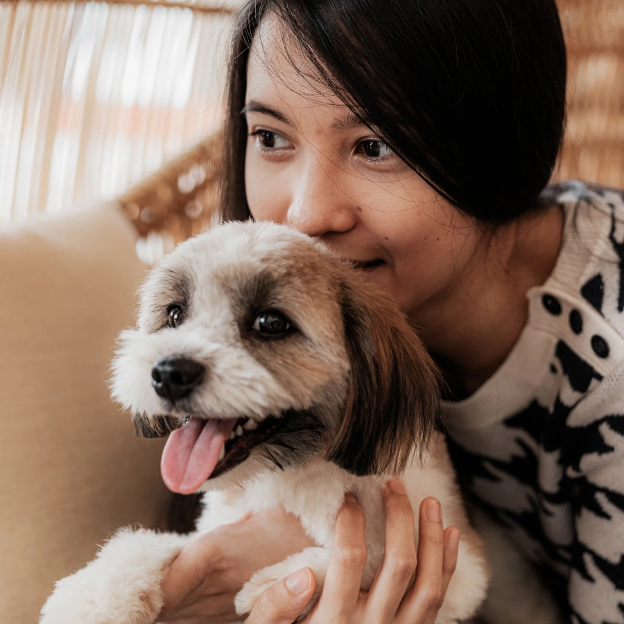 A woman in a patterned top holds a small fluffy dog close to her face, smiling softly as the dog licks its tongue.