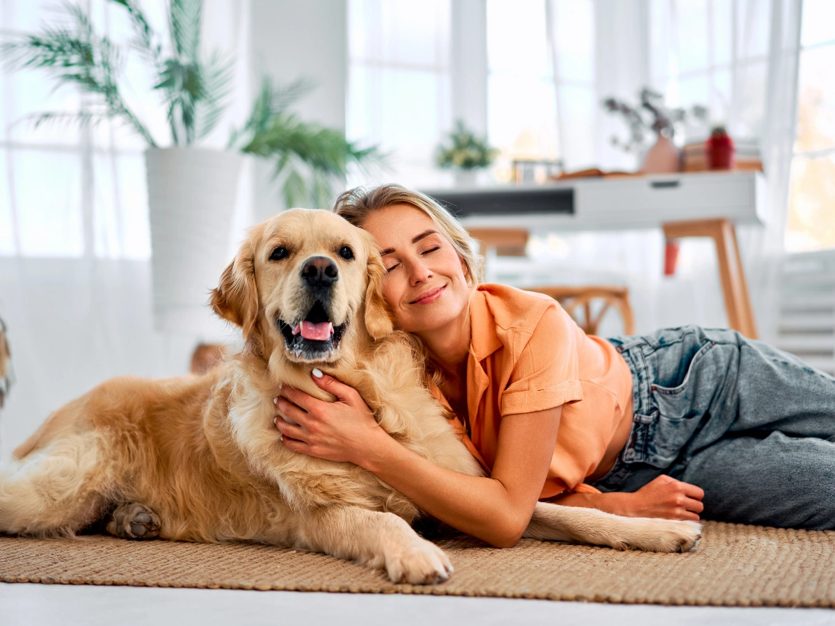A person in an orange shirt smiles while hugging a golden retriever lying on a beige rug in a bright, modern living room with large windows and potted plants.