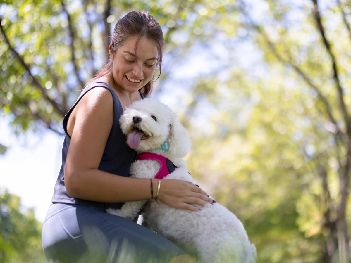 A person in a black tank top and light leggings sits outdoors in a park, gently holding a small white dog with a pink collar that licks its tongue.
