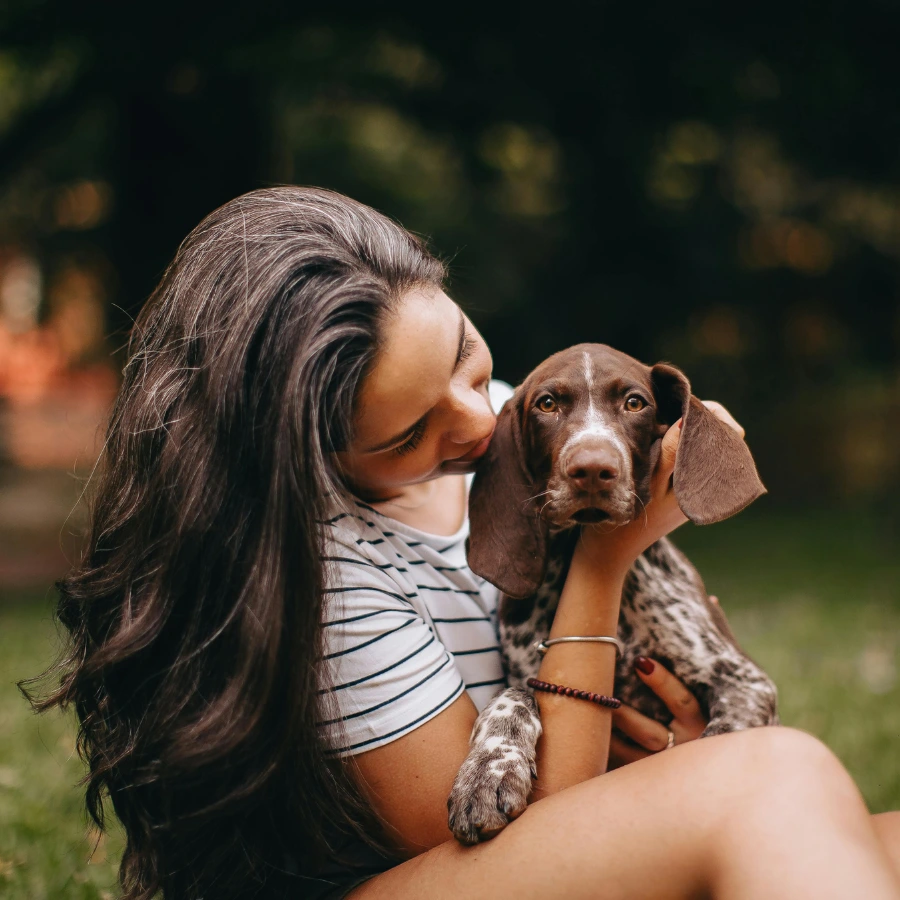 A person with long dark hair gently cuddles a brown-and-white dog outdoors, leaning in to nuzzle the dog’s face as the background blurs into green and warm lights.