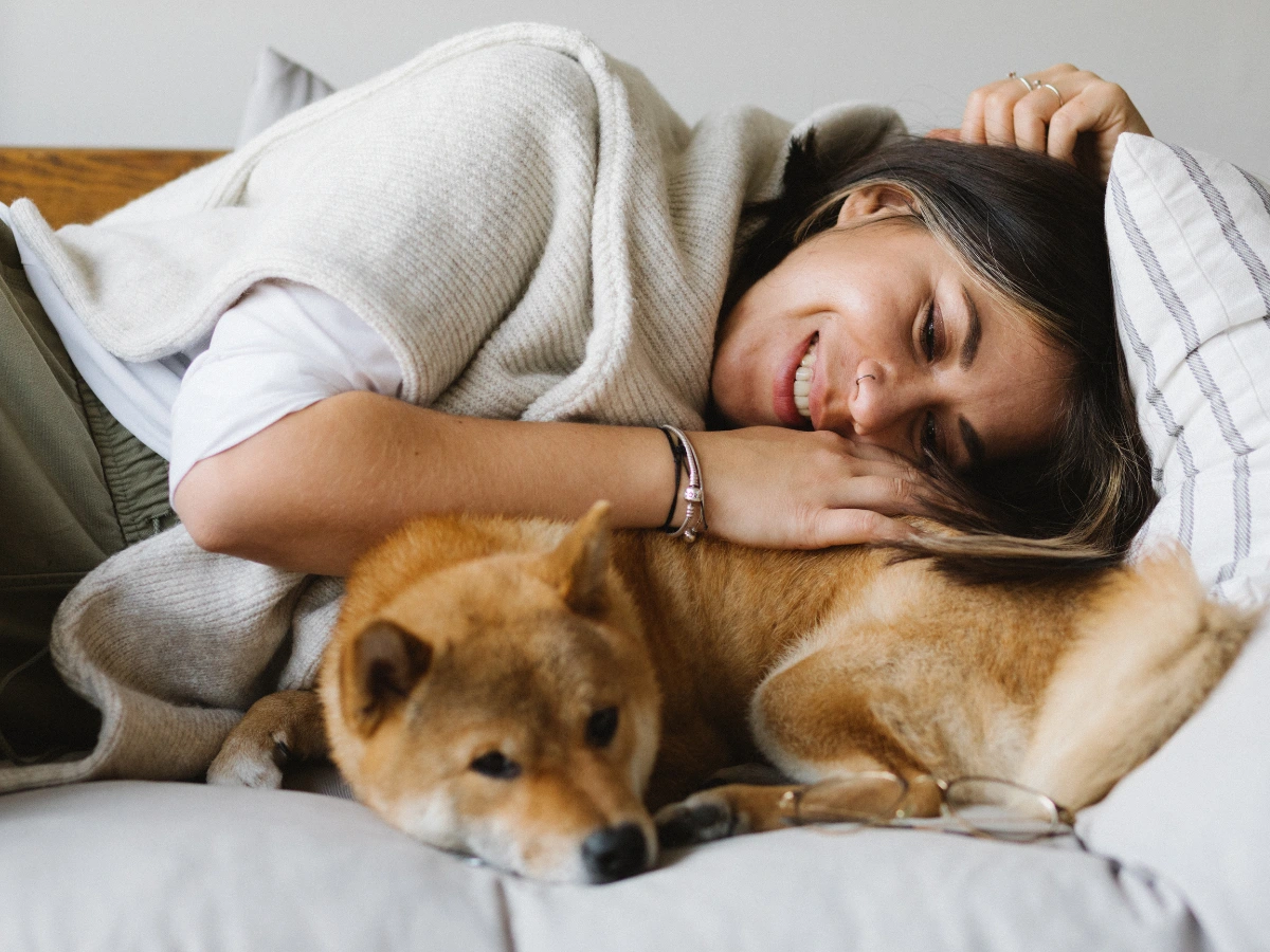 A person sleeping on a sofa with a gray blanket, cuddling a small tan dog lying on the couch beside them.